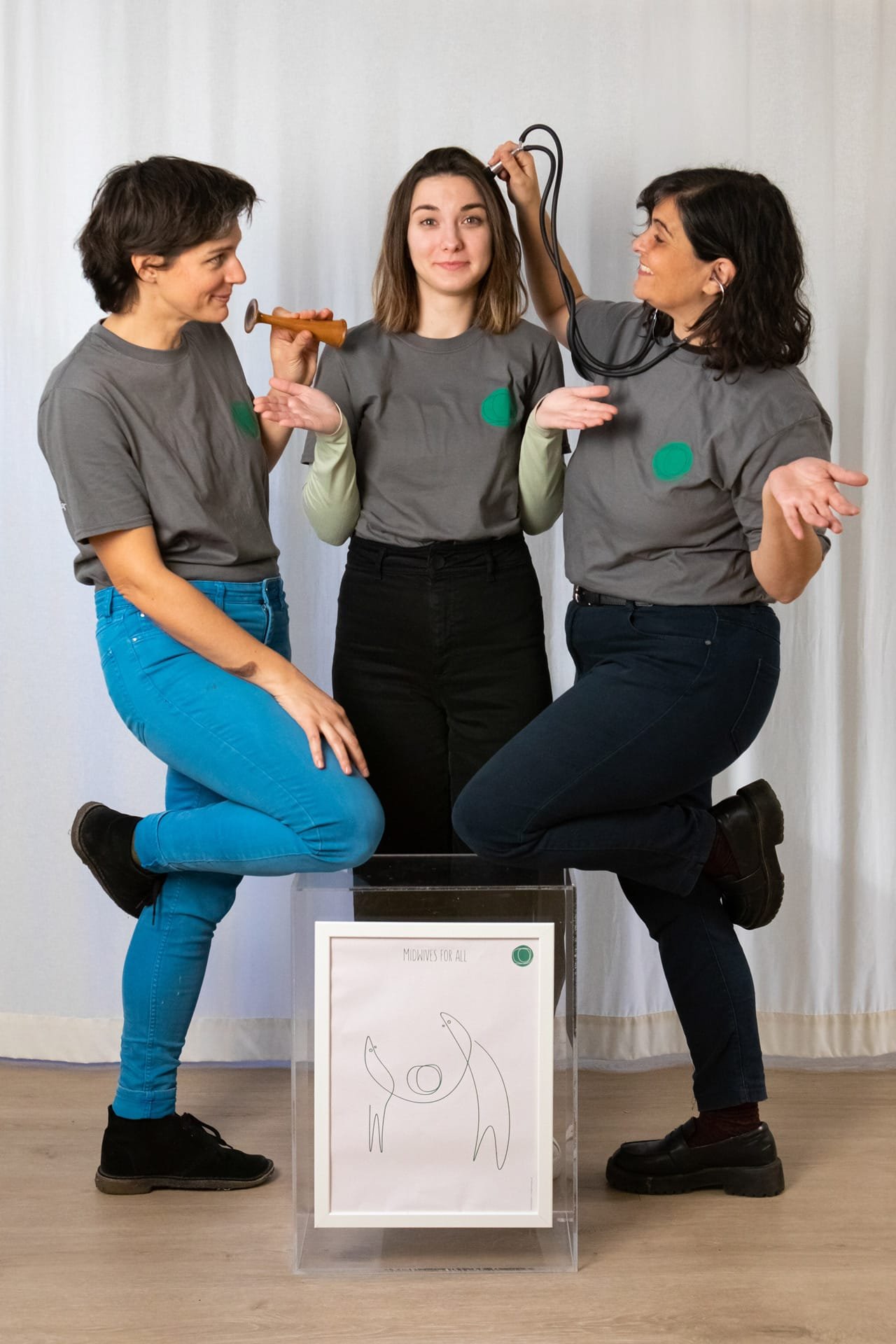 Three young midwives playing around with their medical apparatus, around a framed MFA poster